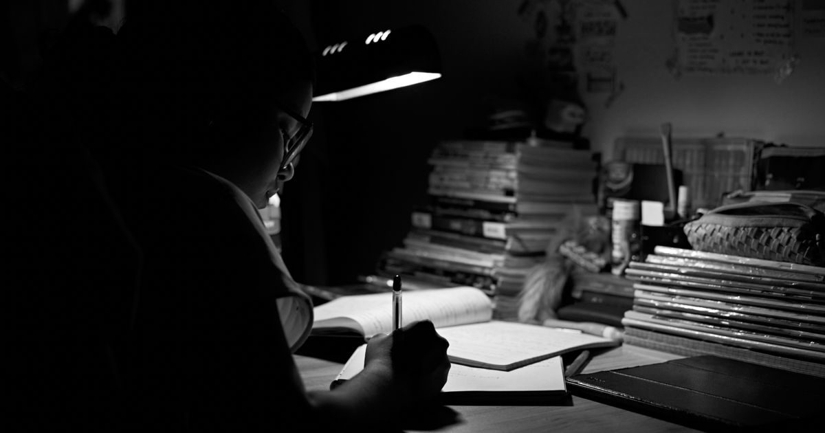 A student intensely studying under a desk lamp, surrounded by books, in a black and white setting.